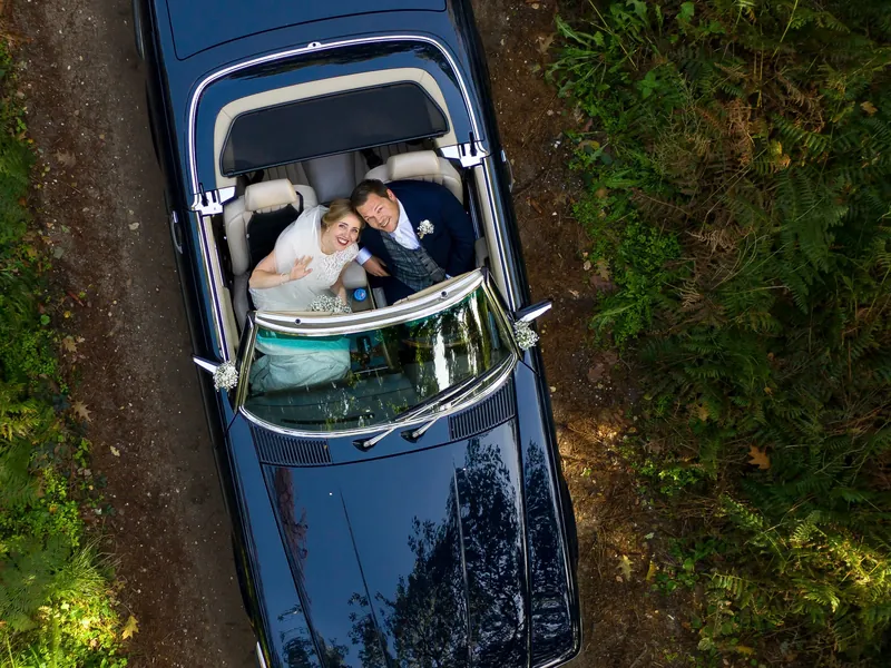Dronefoto van een bruidspaar in een blauwe cabrio op een bospad, ze kijken omhoog naar de camera