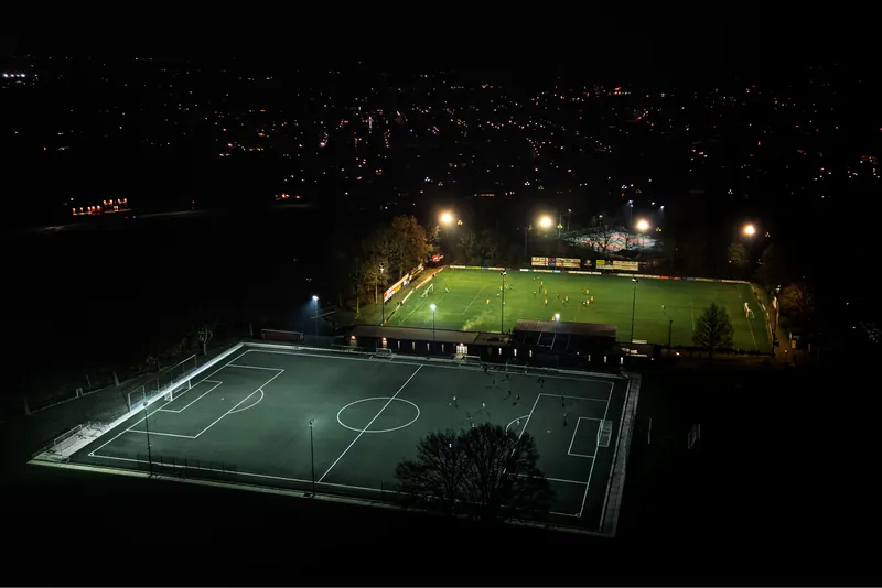 Voetbalveld bij avond vanuit de lucht, verlicht door lichtmasten